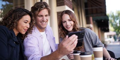 Two female, one male, Spectrum Mobile customers looking at a smartphone smiling while sitting at outdoor cafe